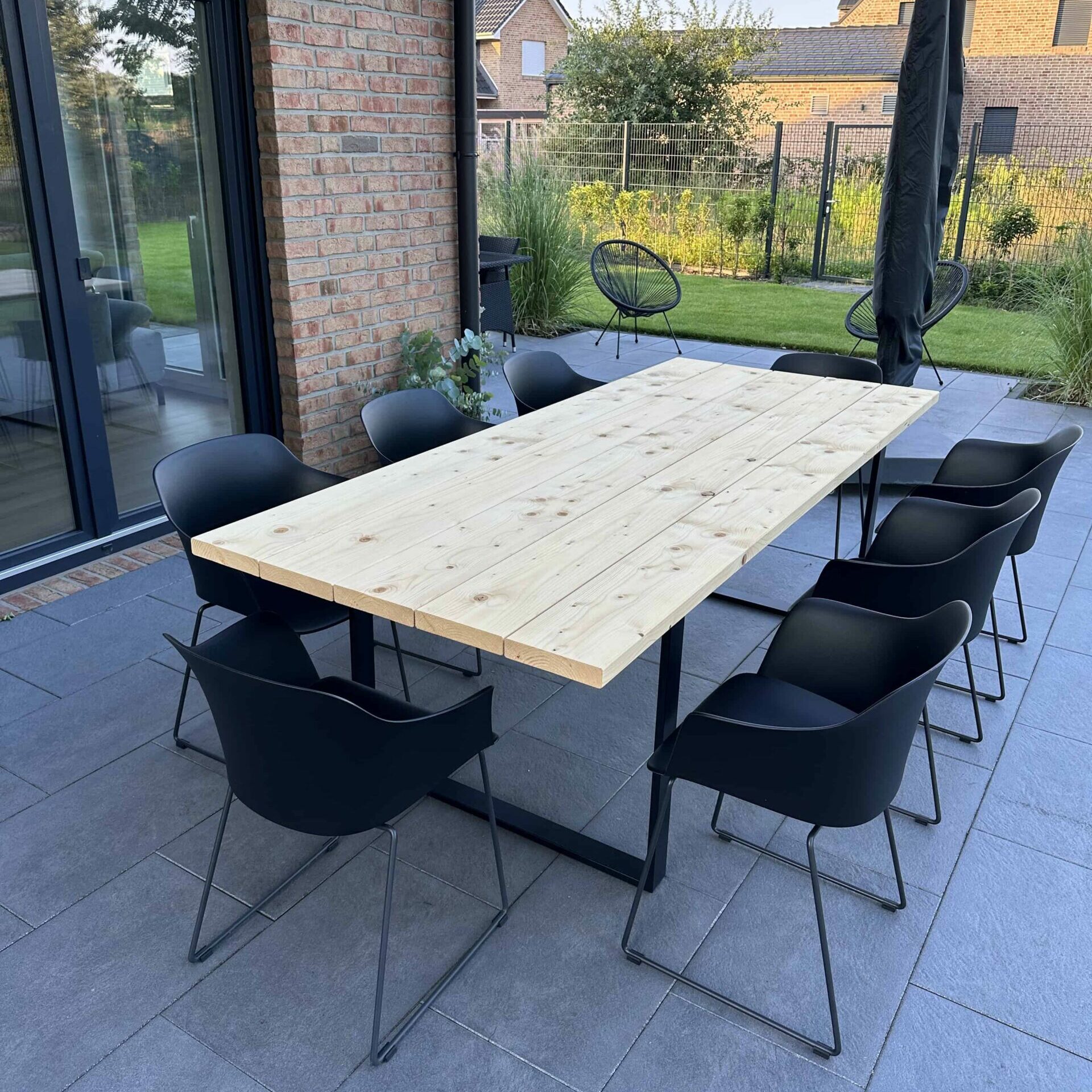 Gartentisch aus hellem Holz mit schwarzen Tischkufen und acht schwarzen Stühlen auf Terrasse
