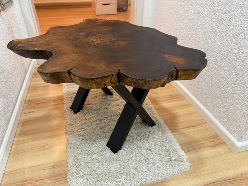 Fertig montierter Unikat-Tisch aus geölter Baumscheibe mit X-Tischbeinen auf Teppich im Innenraum